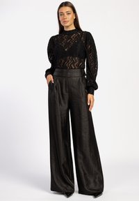 Schwarzes Spitzentop mit Zickzackmuster und Puffärmeln, kombiniert mit hochgeschnittenen, weit geschnittenen schwarzen Hosen aus strukturiertem Stoff.