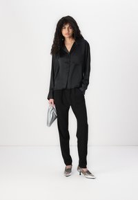 Zwarte satijnen overhemd met knopen, in ontspannen pasvorm, gecombineerd met zwarte broek. Model houdt een zilveren clutch vast; schoenen hebben metalen accenten.