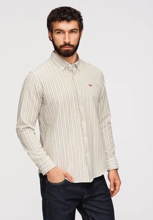 Dorosły mężczyzna z kręconymi włosami i brodą, ubrany w beżową i białą koszulę w pionowe paski na guziki oraz ciemne jeansy, stojący z jedną ręką w kieszeni.