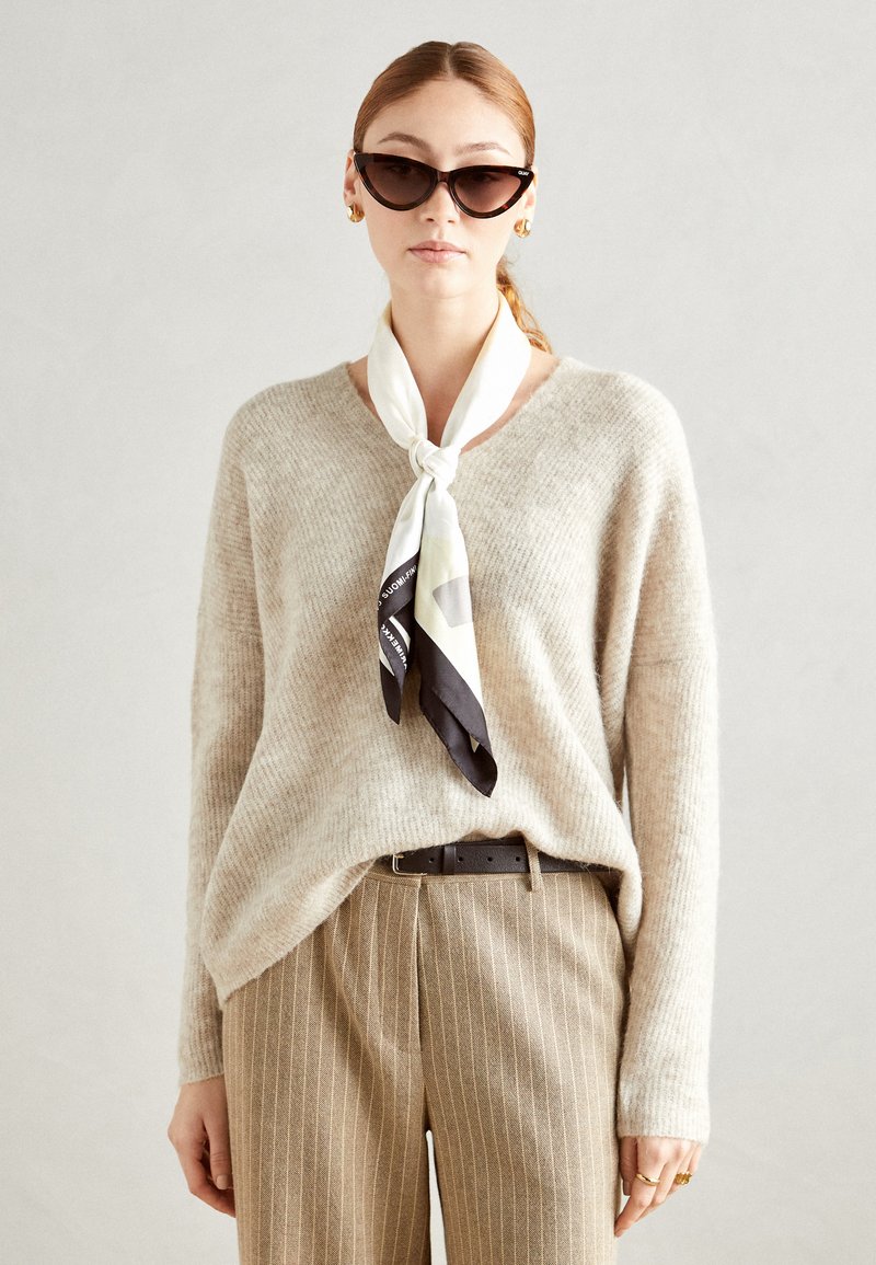 Beige knit sweater, v-neck style, paired with pinstriped beige pants and a multi-colored silk scarf tied at the neck. Cat-eye sunglasses worn.