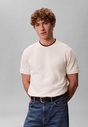 Junger Mann mit lockigem Haar, der ein schlichtes weißes kurzärmeliges Hemd und blaue Jeans trägt, steht vor einem grauen Hintergrund mit den Händen hinter dem Rücken.