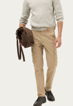 Mann trägt beige Cargohose und grauen Pullover, hält eine braune Schultertasche und steht vor einfarbigem Hintergrund.