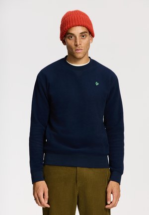 Marineblauwe sweatshirt van een katoenmix, met een groene logo op de linkerborst. Man draagt een rode gebreide beanie en lichtbruine ribfluwelen broek.