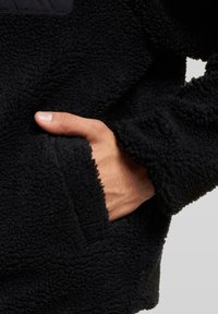 Mustast fliisist kootud pullover, millel on tekstuuriga kangas. Omab külgtaskut ja pehmet, tuhina pinda. Lähedalt vaade näitab kätt taskus.