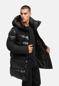 Svart puffjacka med glänsande, vadderat tyg; har en huva, orange foder och framfickor. Modell som bär en svart skjorta under.