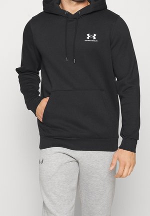 Mężczyzna ubrany w czarną bluzę z kapturem Under Armour z przednią kieszenią i jasnoszare spodnie dresowe, stojący na tle jednolitego jasnego tła.