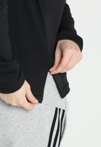 Sweatshirt noir à manches longues, avec une fermeture éclair latérale et un panneau texturé contrastant. Associé à un pantalon de survêtement gris avec des rayures noires.