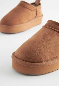 Barna velúr slip-on cipők kerek orral és párnázott talppal. Puha textúrával rendelkeznek, és minimális varrási részletek találhatóak a szegély mentén.