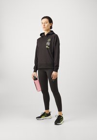 Schwarzer Kapuzenpullover mit grafikdesigns, kombiniert mit schwarzen Leggings. Schwarze Sneakers mit grünen Akzenten. Hält eine pinke Wasserflasche.