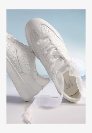 Baskets blanches avec texture en points en relief, semelles épaisses, et larges lacets en ruban transparent, posées sur un fond dégradé bleu et blanc.