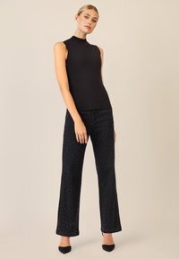 Top nero senza maniche con collo alto; abbinato a jeans scuri con accenti di cucitura bianca sparsi. Modella con scarpe nere a punta.