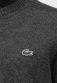 Σκούρο γκρι πλεκτό ύφασμα με μικρό κεντημένο λογότυπο κροκόδειλου σε λευκό, μαύρο και κόκκινο κοντά στη γραμμή του λαιμού.