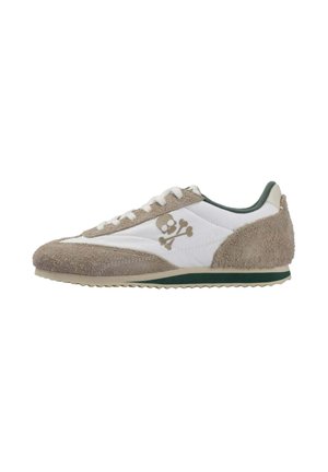 Zapatilla deportiva de ante blanca y beige con un logo de calavera y tibias cruzadas en el lateral y un detalle verde en la suela.