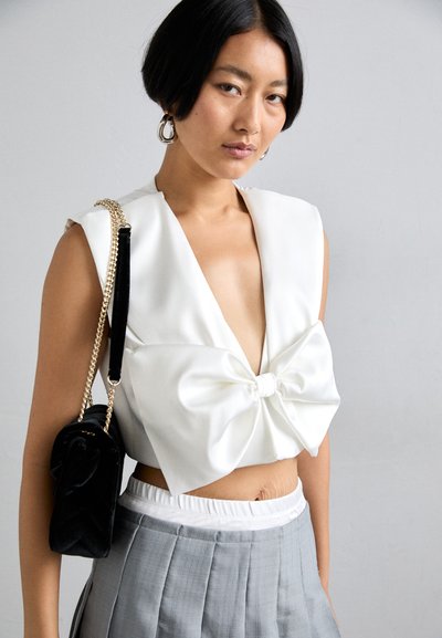 Biały satynowy crop top z głębokim dekoltem w kształcie litery V i dużą kokardą z przodu, zestawiony z szarymi plisowanymi spodniami o wysokim stanie i czarną torbą.