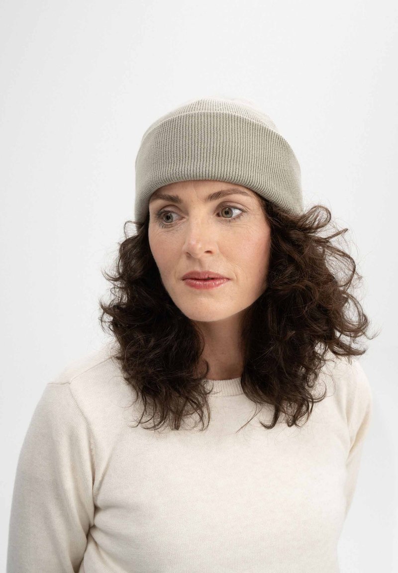 Hellgraue, gerippte Strickmütze, die auf dunklem, gewelltem Haar liegt; kombiniert mit einem cremefarbenen Pullover, der einen runden Halsausschnitt und eine weiche Textur hat.