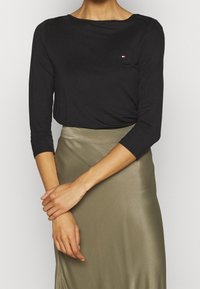 Una modella sta in piedi con le braccia incrociate, indossando un top a maniche lunghe nero e una gonna di seta verde oliva, creando un look elegante e alla moda.