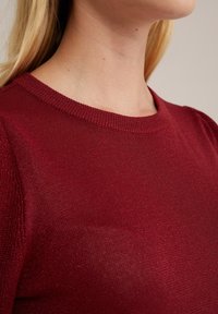 Raudonas megztinis su apvaliu kaklu, turintis subtilų blizgesį ir lygų paviršių. Dizainas apima šiek tiek pūstus rankovės.