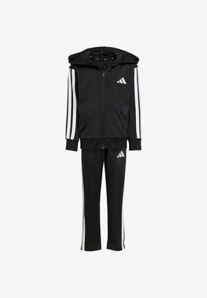 Dwójka dzieci stoi obok siebie w czarnych dresach Adidas z białymi paskami; dziewczynka ma na sobie różową koszulkę, oboje się uśmiechają.