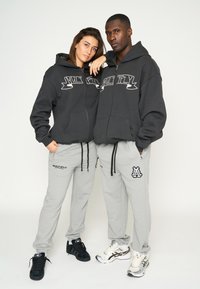 Sudaderas gris oscuro con el texto "MULTIPLY", combinadas con pantalones de chándal gris claro que tienen logotipos. Zapatillas negras y plateadas complementan el atuendo.