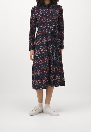 Vrouw draagt een marineblauwe midi-jurk met bloemenprint, lange mouwen, knopen aan de voorkant, tailleceintuur, witte sokken en witte sneakers, staand op een effen achtergrond.