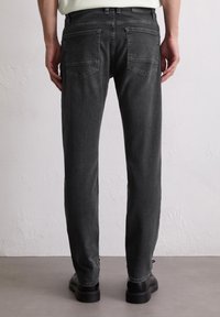 Persoon in slim-fit zwarte spijkerbroek met achterzakken en zwarte schoenen, staand tegen een effen witte textuurmuur.