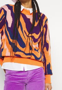 Suéter de punto en tonos naranja, rosa y purpura con un patrón de remolino; presenta cuello y puños de canalé. Combinado con pantalones de pana morados.
