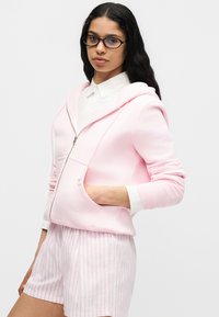 Femme aux longs cheveux foncés et lunettes, portant un sweat à capuche rose clair zippé sur un t-shirt blanc et un short rose rayé, debout avec la main dans la poche.