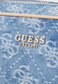 Dongerwallet med lys blå farge, teksturert mønster og blank sølv "GUESS EST 1981" logo. Glidelås lukking med dekorative detaljer.