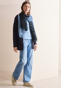 En blå og sort tørklæde svøber sig omkring en lyseblå top, parret med lyseblå jeans og beige sneakers, der har en afslappet pasform og bløde teksturer.
