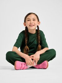 Fille aux cheveux bruns tressés, assise en tailleur, portant une tenue vert foncé et des baskets roses vives à scratch, souriante sur un fond uni.
