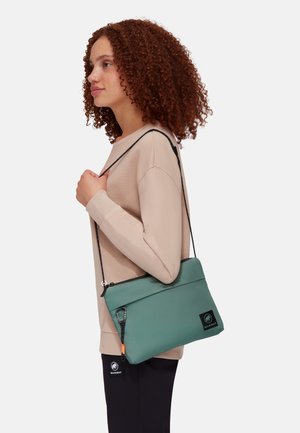 Grüne strukturierte Schultertasche mit Reißverschluss, ausgestattet mit einem verstellbaren schwarzen Tragegurt und einem Logo-Patch, über einem beigen Pullover getragen.