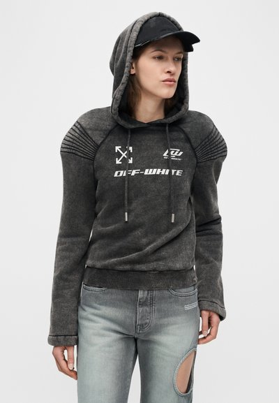 Γυναίκα που φοράει μια ξεθωριασμένη μαύρη hoodie Off-White με επενδυμένους ώμους και μαύρο καπέλο, συνδυασμένη με ανοιχτό μπλε τζιν με στρογγυλό άνοιγμα στο γόνατο.