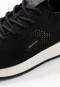 Black athletic shoe featuring a knitted upper with mesh panels, lace-up design, and a white cushioned sole. GANT logo in gold on the side.