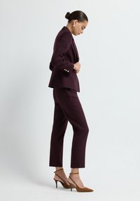 Blazer ajusté bordeaux et pantalon fuselé en tissu lisse, associés à des talons en daim marron avec des brides aux chevilles.