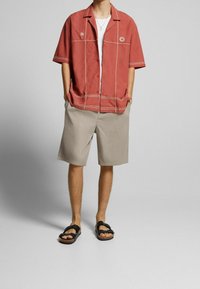 Camicia rossa a maniche corte con bordature bianche, pantaloni corti beige e sandali neri. La camicia presenta ricami di palme e sole.