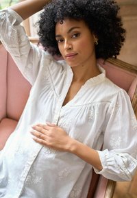 Hvid bluse med blomsterbroderi, flæset halsudskæring og sammenskruede ærmer. Modellen har krøllet hår og sidder ned, mens hun blidt rører ved sin mave.