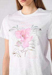 Hvit bomull T-skjorte med et rosa blomstertrykk, sølvdetaljer og teksten "DON'T WORRY BE HAPPY" i pastellfarger. Korte ermer.