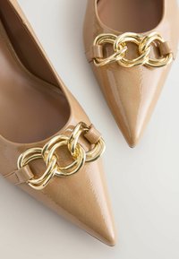 Des chaussures à talons hauts beiges avec des pointes et un détail de chaîne dorée décoratif sur l'empeigne, sur un fond neutre.