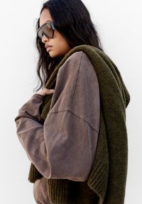 Übergroßer brauner Pullover aus strukturiertem Stoff, getragen mit einem dunkelgrünen Wollschal, der über die Schulter drapiert ist. Dicke Rippenakzente am Schal.