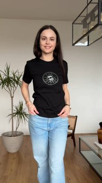 Νεαρή γυναίκα χαμογελάει, φοράει μαύρο γραφικό μπλουζάκι και ανοιχτό μπλε τζιν, στέκεται σε ένα δωμάτιο με φυτά, μια καρέκλα και ένα γυάλινο τραπεζάκι καφέ.