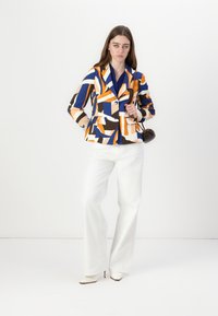 Blazer multicolore con motivi geometrici in blu, arancione e crema; presenta un colletto a banda, chiusura con un unico bottone e tasche frontali.