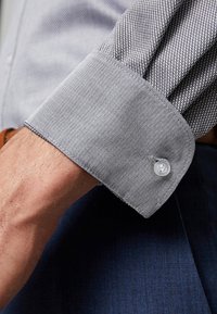 Gros plan sur le poignet d'une personne portant une chemise grise à motif chevrons, avec le poignet boutonné, associée à un pantalon bleu foncé.
