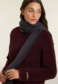 Sciarpa lavorata a maglia grigia scura drappeggiata attorno al collo, abbinata a una giacca in pile bordeaux con texture e un piccolo logo sulla manica.