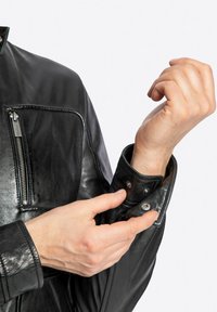 Chaqueta de cuero negro con un bolsillo en el pecho con cremallera, cierres a presión en los puños y textura suave; manos ajustando el puño visibles.