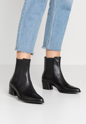 Bottines en cuir noir texturé avec talons carrés portées avec un jean court bleu clair sur un fond neutre.