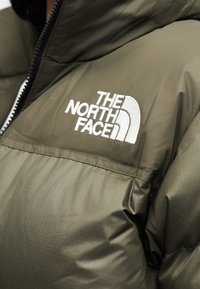 Πούπουλο μπουφάν North Face σε χακί πράσινο με λευκό κεντητό λογότυπο στο στήθος κοντά σε μαύρο φερμουάρ.