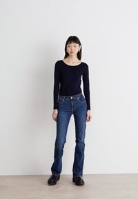 Navy long-sleeve top paired with fitted dark blue jeans. Model wears black chunky shoes, standing on a light floor against a plain background.