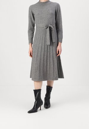 Robe tricotée grise avec des manches longues, un corsage ajusté et une jupe plissée. Comprend une ceinture à nouer. Associée à des bottes noires à talons hauts et à bouts pointus.