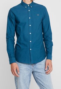 Camisa de color verde azulado hecha de algodón, con cuello abotonado, tapeta frontal con botones blancos y un pequeño logo en el pecho.