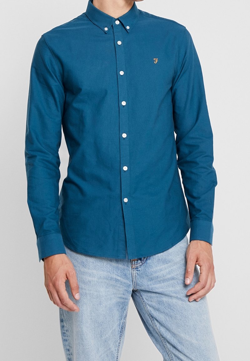 Camisa de color verde azulado hecha de algodón, con cuello abotonado, tapeta frontal con botones blancos y un pequeño logo en el pecho.